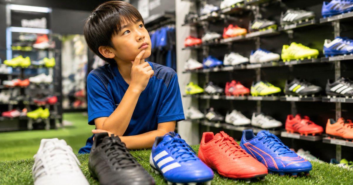スポーツ用品店で複数のサッカースパイクを前に、どれを選ぶか考える少年