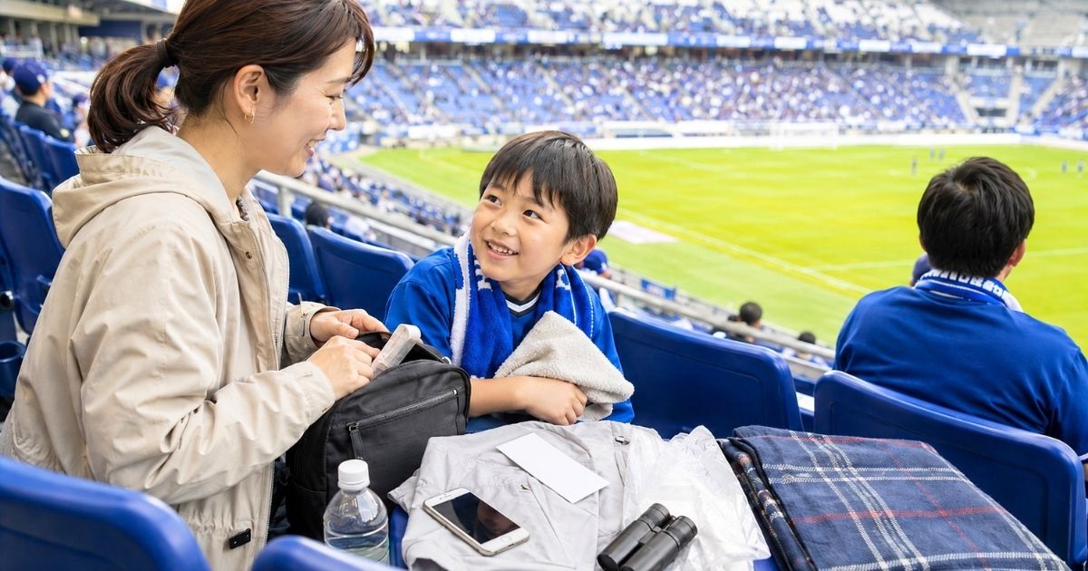 Jリーグ観戦の準備をする親子。スタジアムの座席にバッグ、飲み物、スマホ、双眼鏡、ブランケットなどの持ち物を置いている様子
