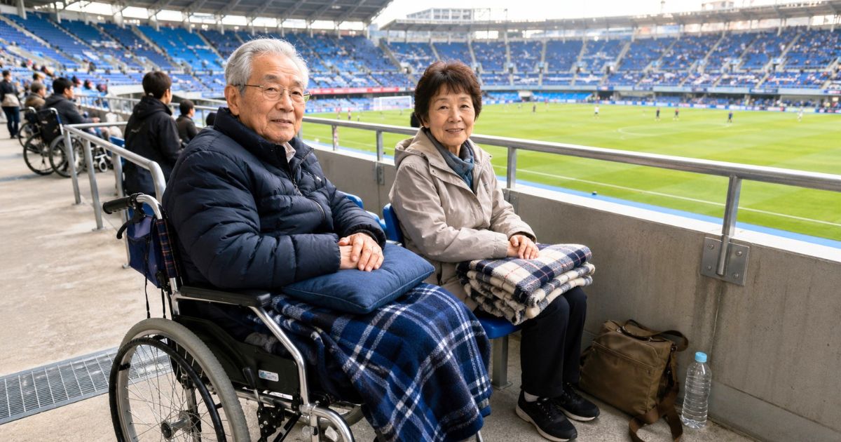 車椅子利用の高齢男性と同伴女性が、ひざ掛けや飲み物を用意してスタジアムの観戦エリアでサッカーを観る様子