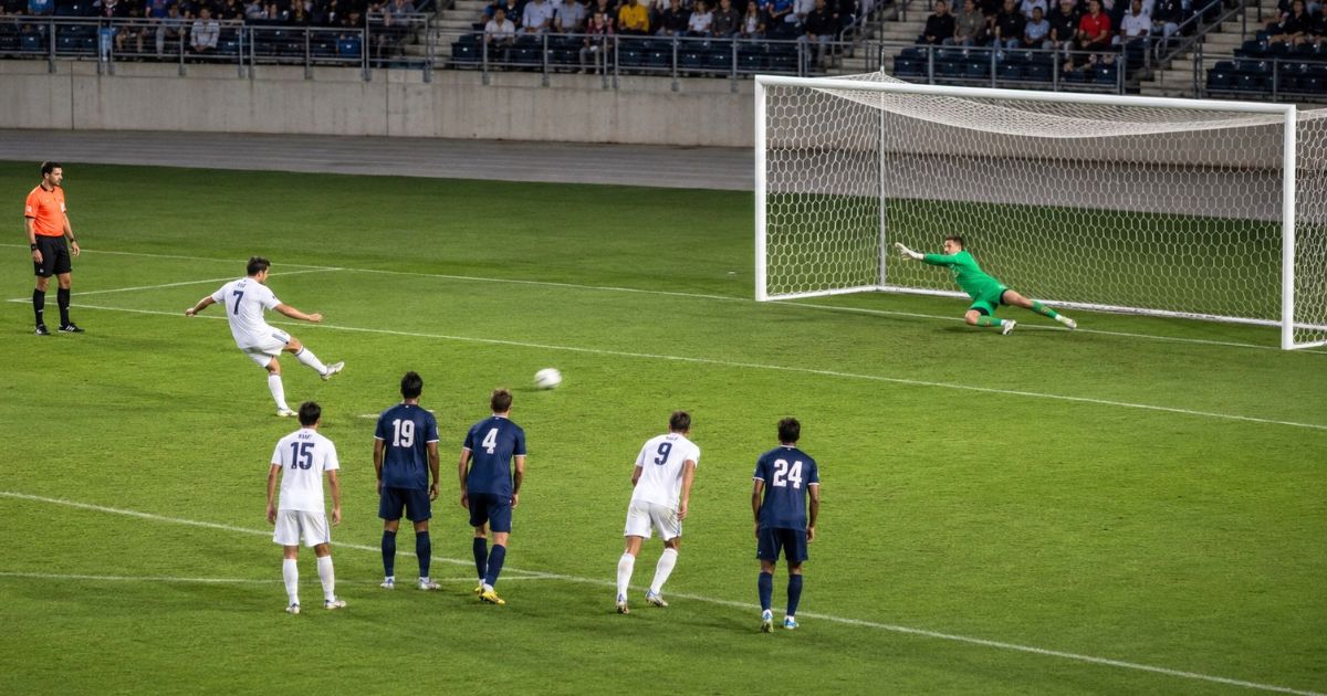 大学サッカーのインカレ予選ラウンドで、選手がPKを蹴、GKがゴール前で横へ飛び、審判と両チームの選手が見守る場面