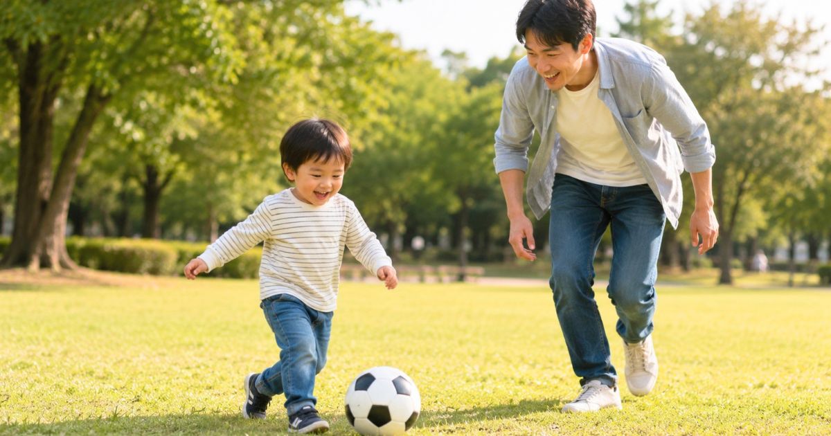 公園の芝生でサッカーボールを追いかけて遊ぶ3歳くらいの子どもと日本人の父親