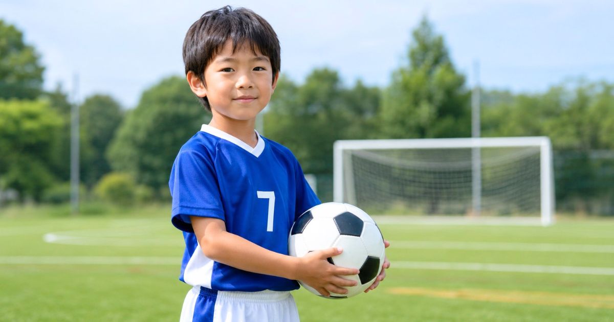 サッカーゴールのあるグラウンドを背景に、ボールを抱えた青いユニフォーム姿の少年サッカー選手