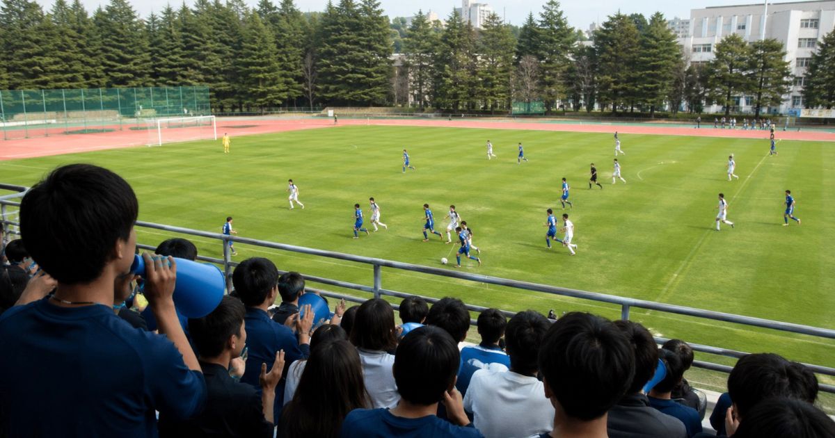 関西学生サッカーリーグ2部・3部プレーオフを応援席から観戦する学生と、ピッチで競り合う大学選手たち