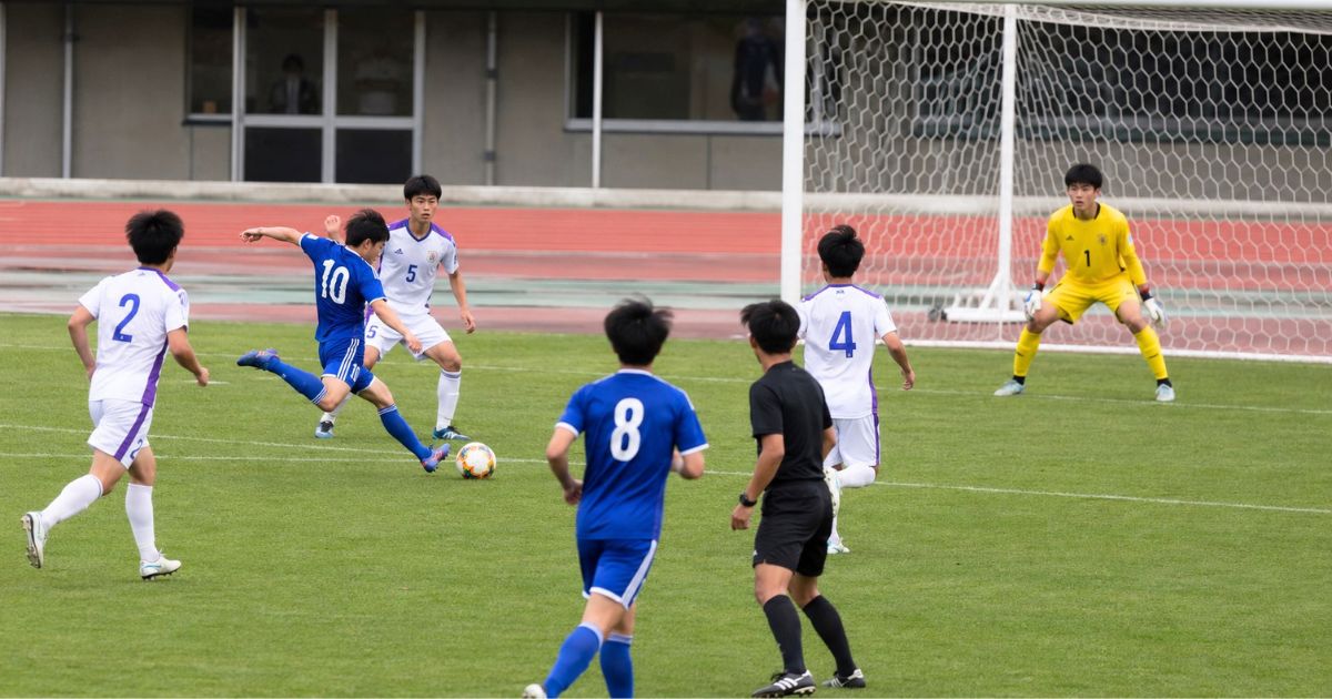 地域リーグの公式戦で、青いユニフォームの選手がシュートを放ち、ゴール前でGKが構え、審判も見守る試合シーン