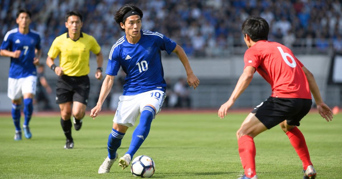 ヘアバンドを着けた選手が、審判の見守るサッカーの試合で相手選手をかわしながらドリブルする様子
