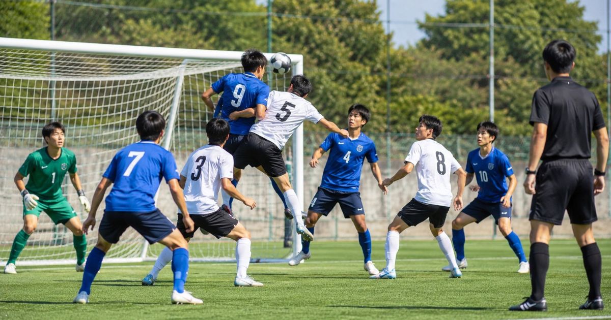 大学サッカーIリーグの試合で、審判が見守る中、背番号入りの選手たちがゴール前で空中のボールを競り合う場面