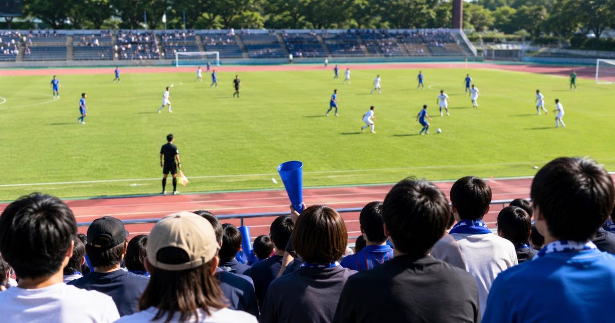 応援席から大学サッカーIリーグの試合を観戦し、青と白のユニフォームの選手を応援する観客