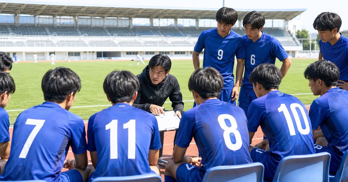 大学サッカーIリーグの試合前、背番号入りの選手たちがベンチで監督から戦術説明を受ける場面