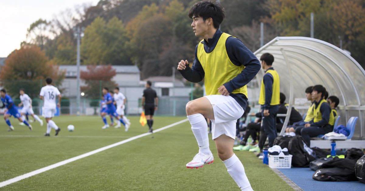 大学サッカーIリーグの試合中、選手交代に備えてベンチ横でウォームアップするビブス姿の控え選手