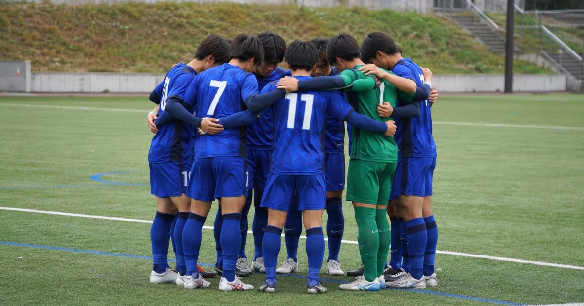 大学サッカーIリーグの試合開始直前、ゴールキーパーを含む選手たちが円陣を組む場面
