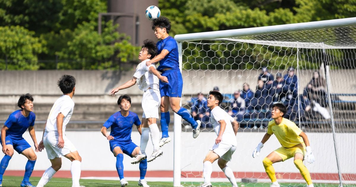 大学サッカーリーグの公式戦で、ゴール前の空中戦でヘディングを競り合う選手たち