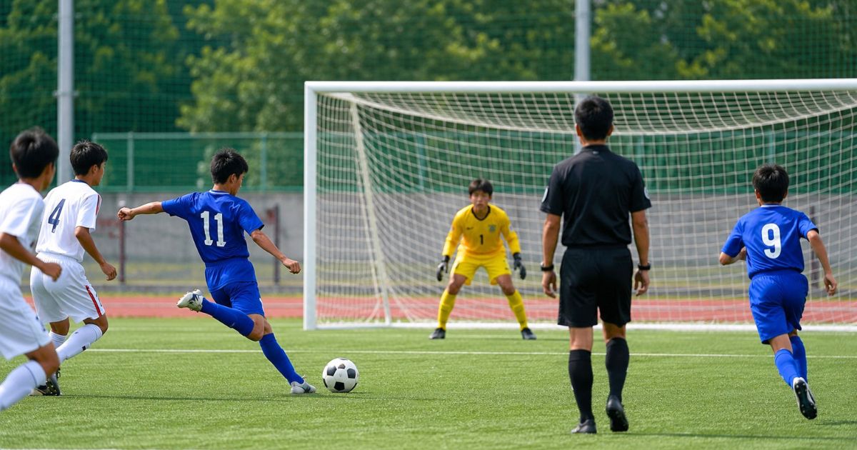 12歳くらいの少年サッカー公式戦で、青いユニフォームの選手がシュートを狙い、ゴール前でキーパーと審判が見守る試合風景