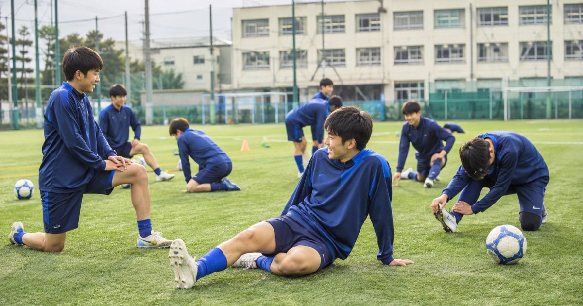 高校サッカー部の練習グラウンドで、選手たちがリラックスしてストレッチする様子