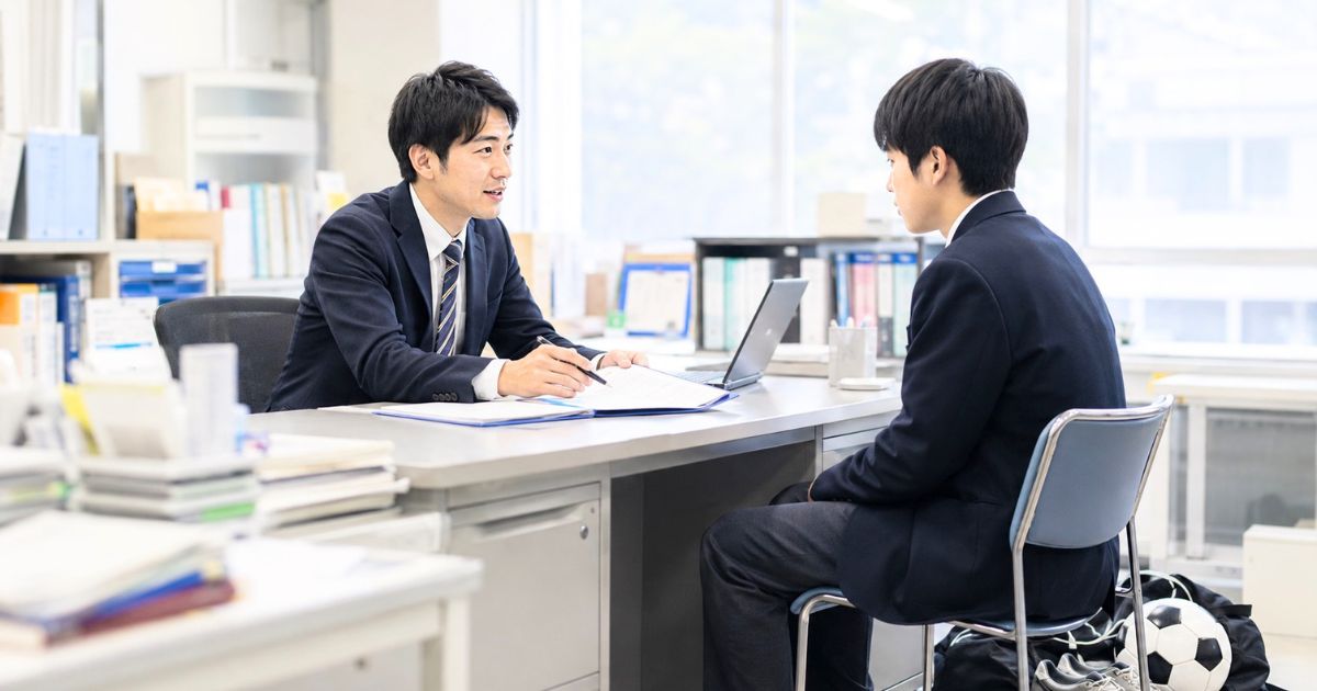 高校の職員室で、大学サッカー部への進学を希望する男子生徒が顧問の先生に相談している様子
