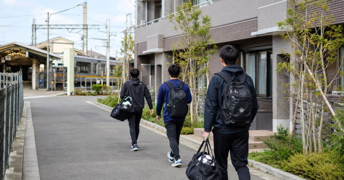 八幡山駅周辺の住宅街をサッカー用具を持って歩く大学生と、駅や線路が見えるアクセス風景
