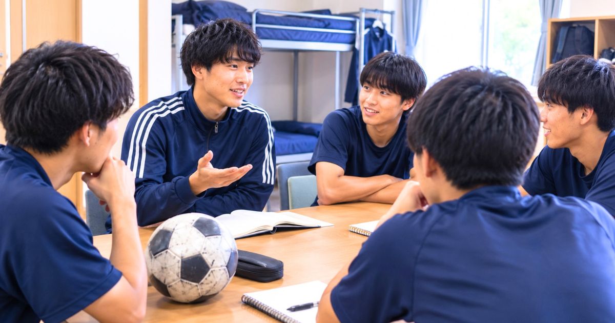 寮の部屋で、サッカー部の寮生活について話し合う日本人大学サッカー部員たちのイメージ