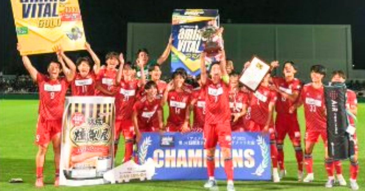 流通経済大学サッカー部の選手たちが優勝トロフィーを掲げ、ピッチ上で喜ぶ集合写真