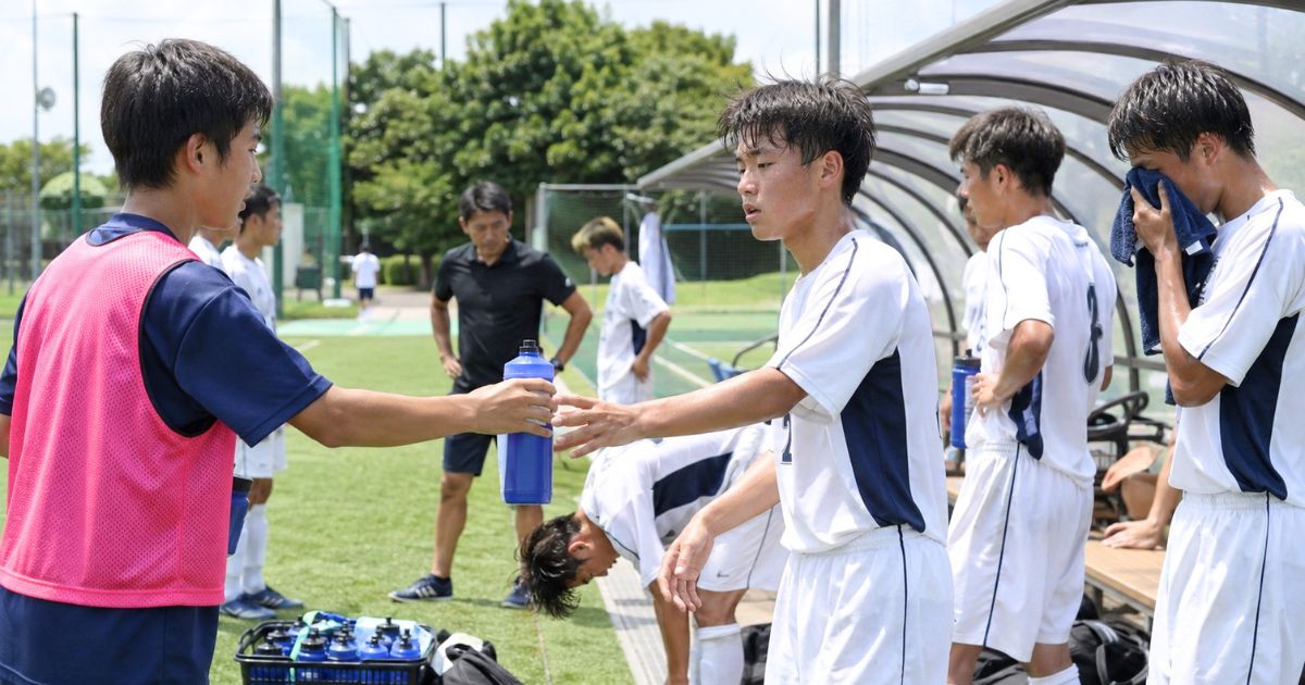 公式戦のハーフタイムに、控え選手が疲れた高校生年代の選手へ給水ボトルを手渡すベンチ前の様子