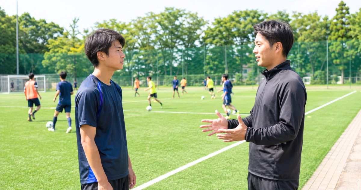 大学サッカーのグラウンドで練習を背景にコーチと話す入部希望者