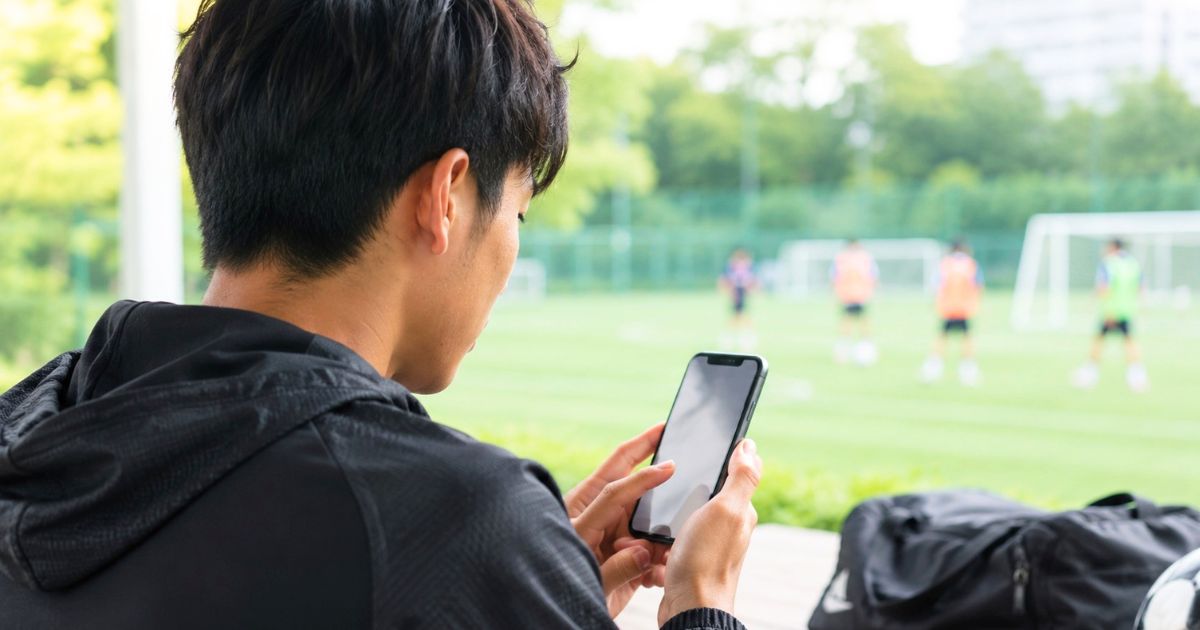 大学サッカーの練習参加を希望する学生が、サッカー場を背景にスマートフォンで申し込みをする様子