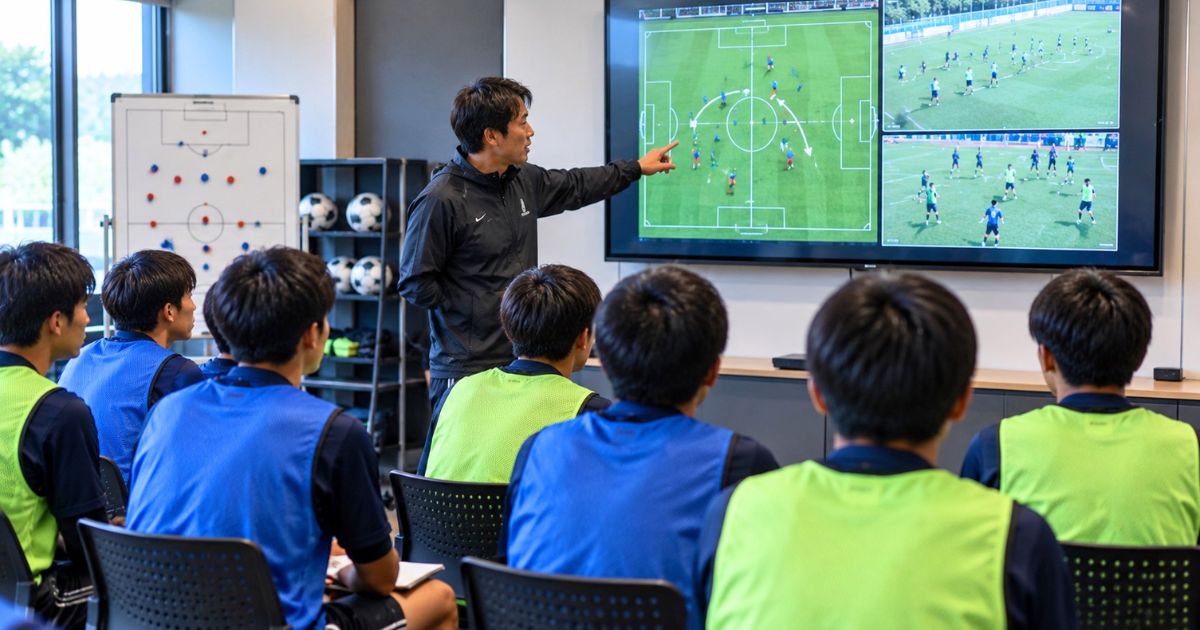 クラブハウスのミーティングルームで、コーチがスクリーンを使って戦術を説明し、トレセン選手たちが聞いている様子