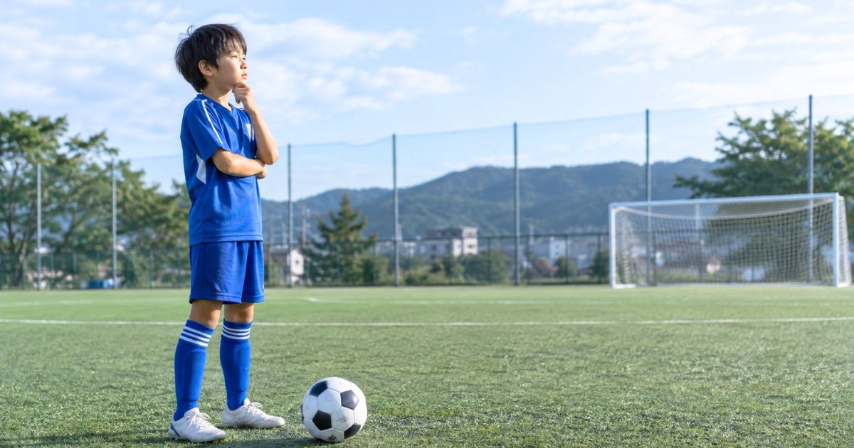 U12年代のサッカー選手がグラウンドでボールのそばに立ち、顎に手を当ててフォーメーションを考えている様子