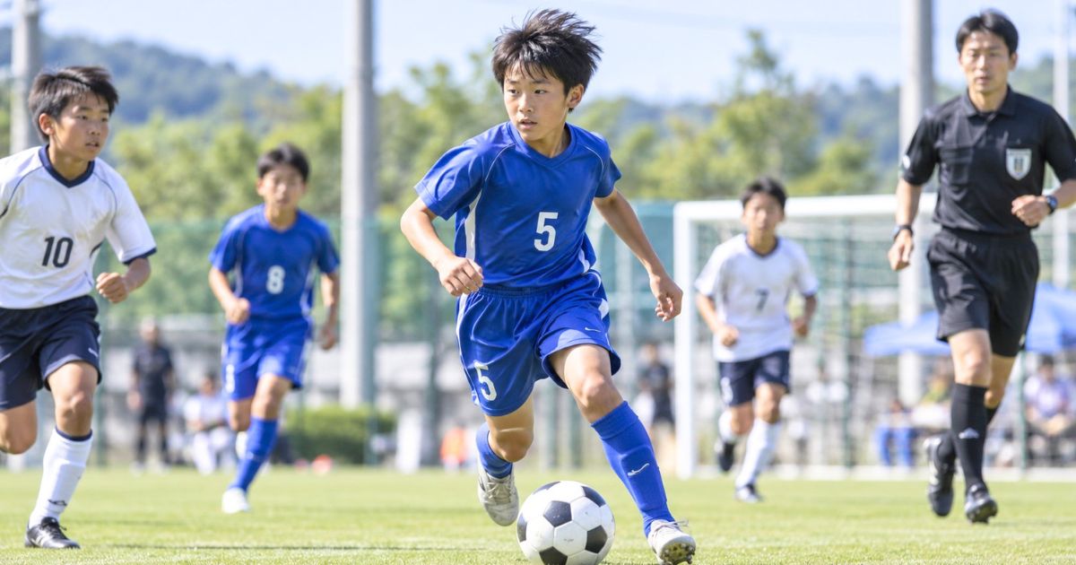 少年サッカーの試合で背番号5の選手がドリブルする様子と審判