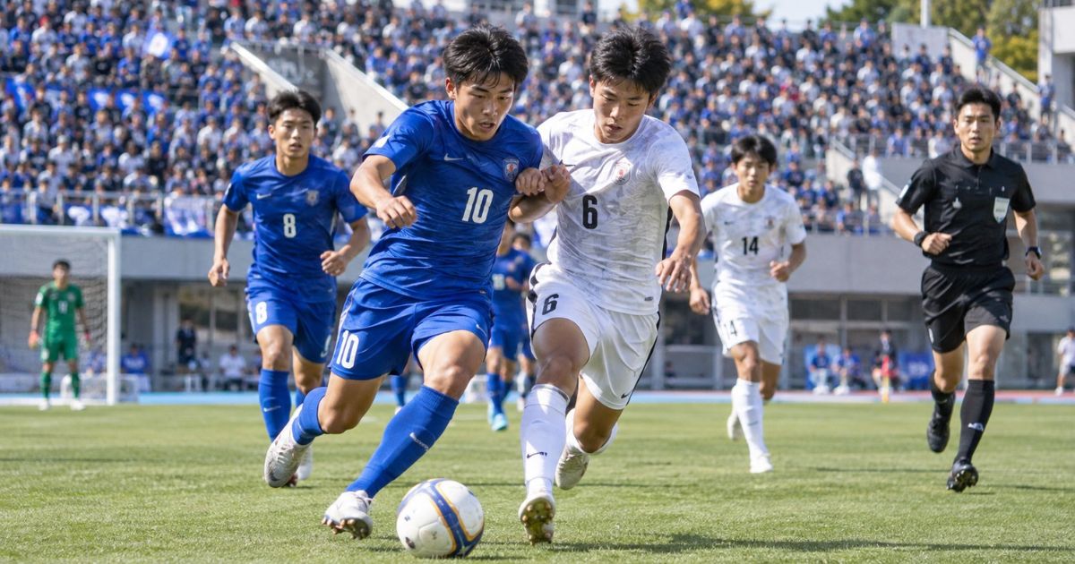 高校サッカーのプレミアリーグをイメージした試合風景。青色の10番と白色の6番の選手がボールを競り合い、審判や満員の応援席が見えるスタジアムで熱戦を繰り広げている。