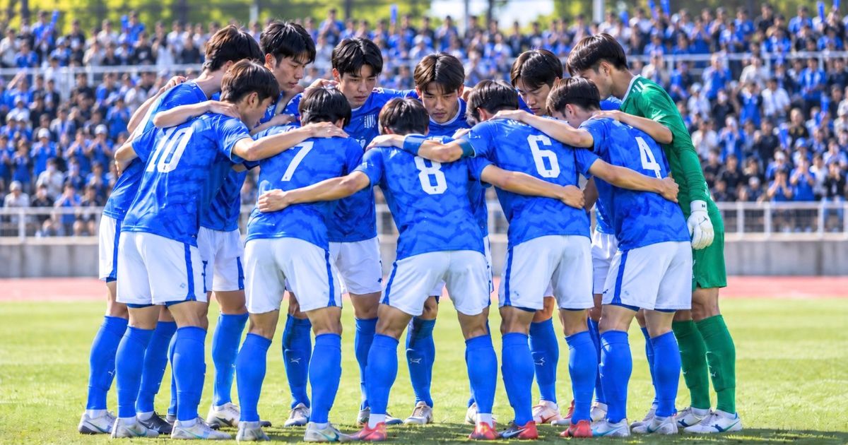 試合前のピッチで円陣を組む高校サッカー選手と、スタンドから声援を送る多くの応援団