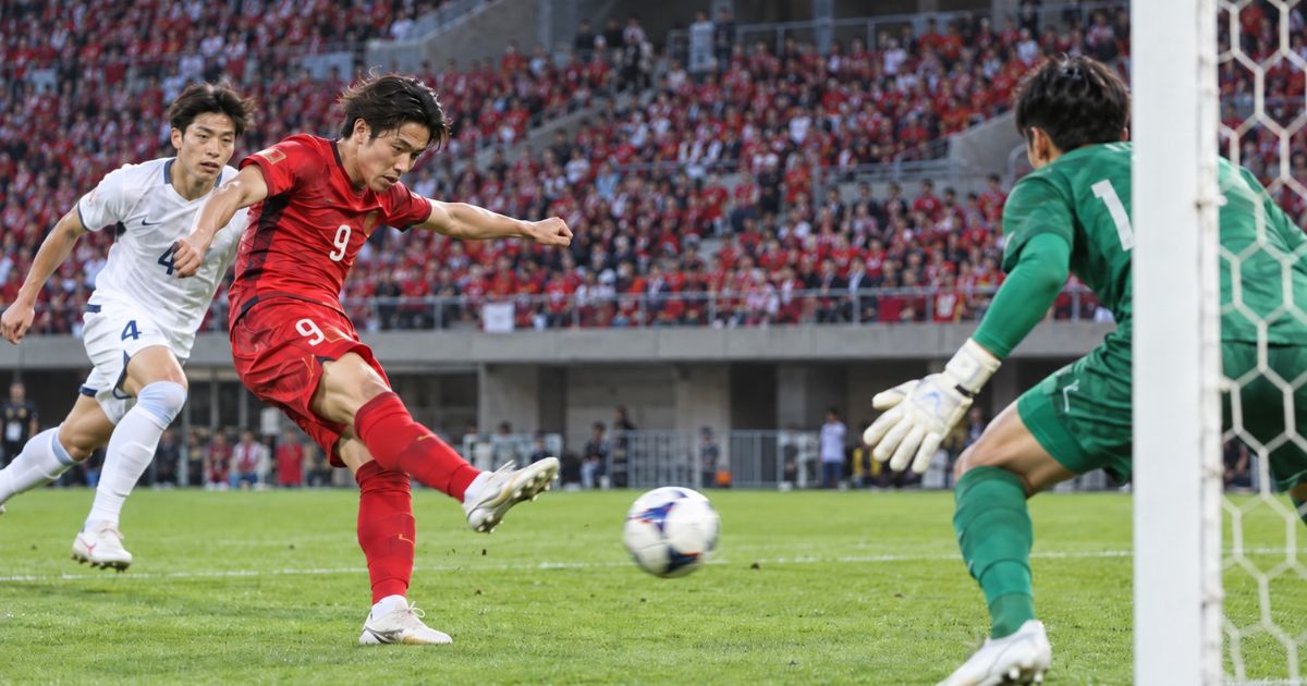 Jリーグの試合をイメージした実写風画像。赤いユニフォームの9番の選手が強烈なシュートを放ち、白いユニフォームの4番が追走。緑のユニフォームのGKがゴール前で構えている。