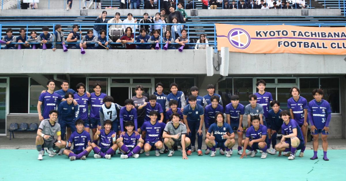 京都橘大学サッカー部の選手たちが紫色のユニフォーム姿で集合し、スタンドの関係者とともに撮影した記念写真。