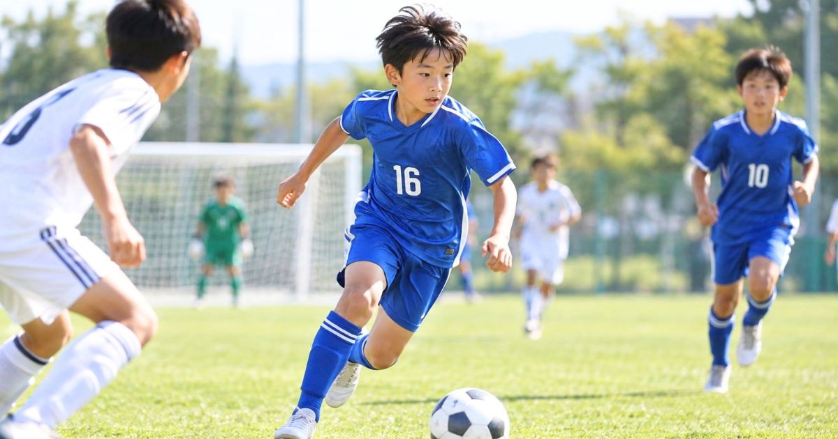背番号16の青いユニフォームを着た少年サッカー選手が、試合中に相手選手へドリブルで仕掛けるシーン