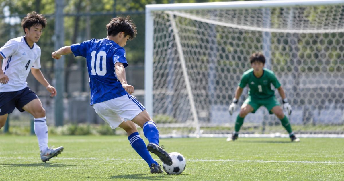 サッカーのシュートモーションを実演する青い10番の選手。ゴール前で白いユニフォームのDFが迫る中、軸足で身体を支え、右足でシュートを打とうとしている。