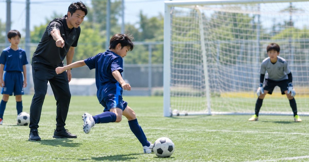 屋外のサッカーグラウンドで、コーチの指導を受けながらシュートモーションの練習をするサッカー少年。ゴールに向かって右足でシュートを打とうとしており、ゴールキーパーと別の少年が見守っている。