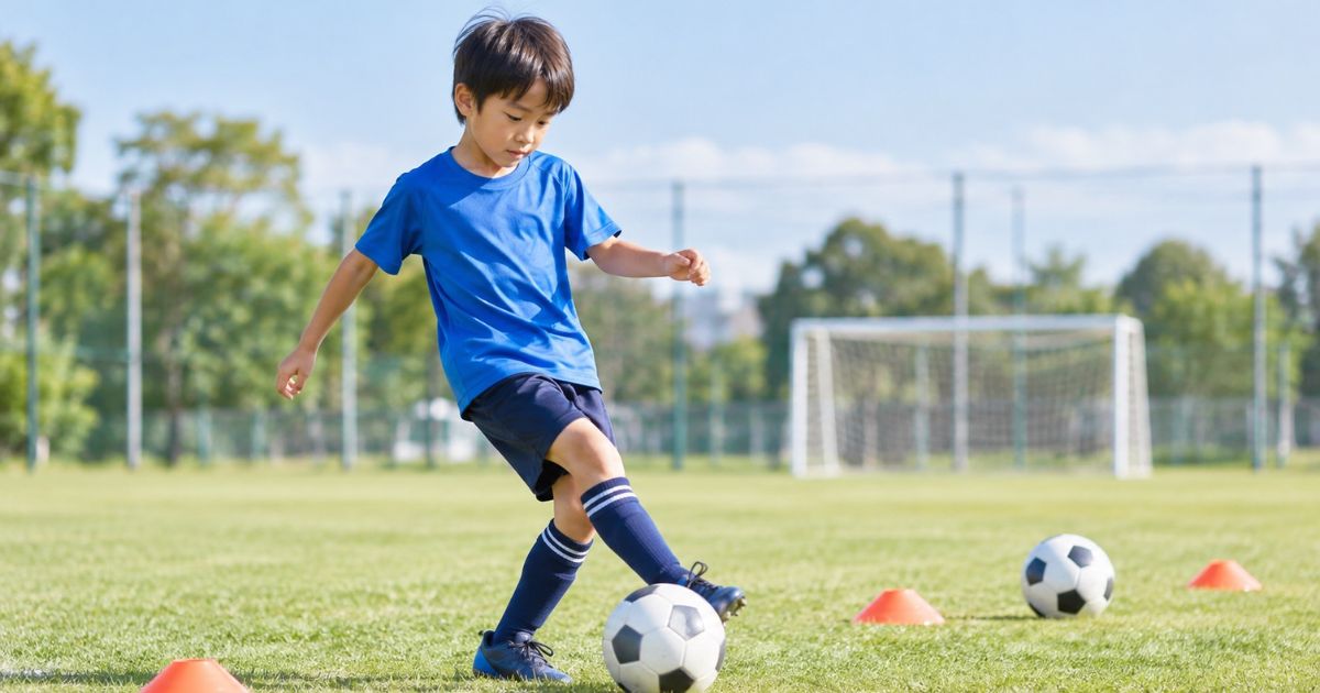 サッカー場で逆足を使ったボールコントロールを練習する小学生の男の子。芝生の上にボールとマーカーコーンを置き、足元のボールを操作している様子。